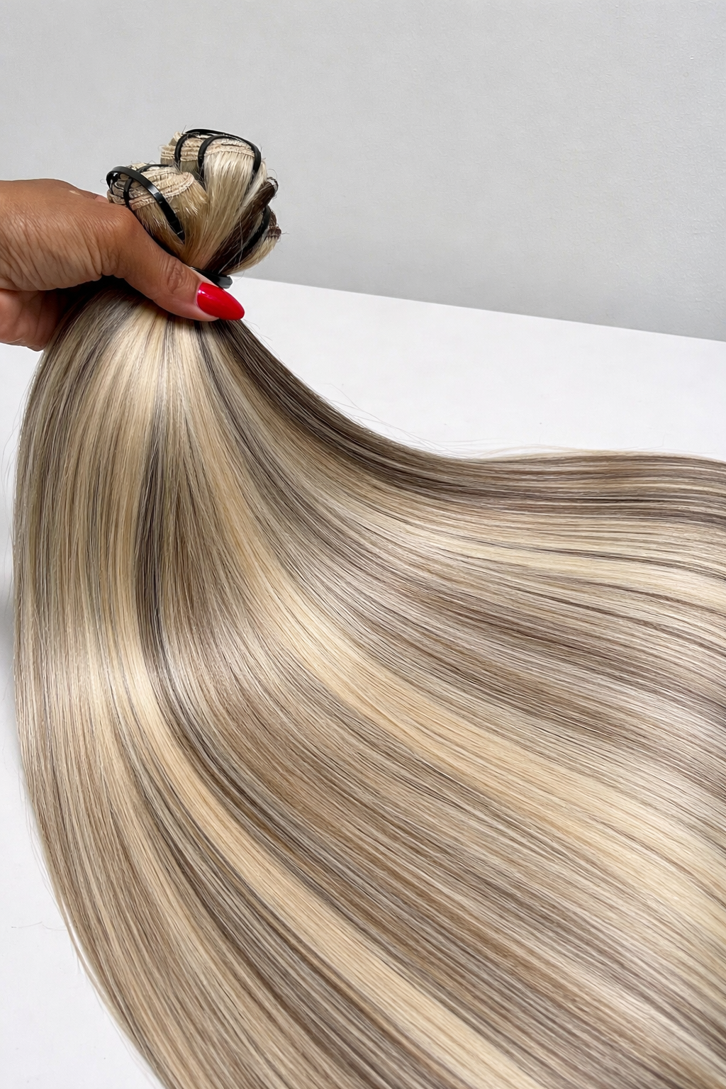 Blonde hair extensions held by a hand with red nail polish on a white background